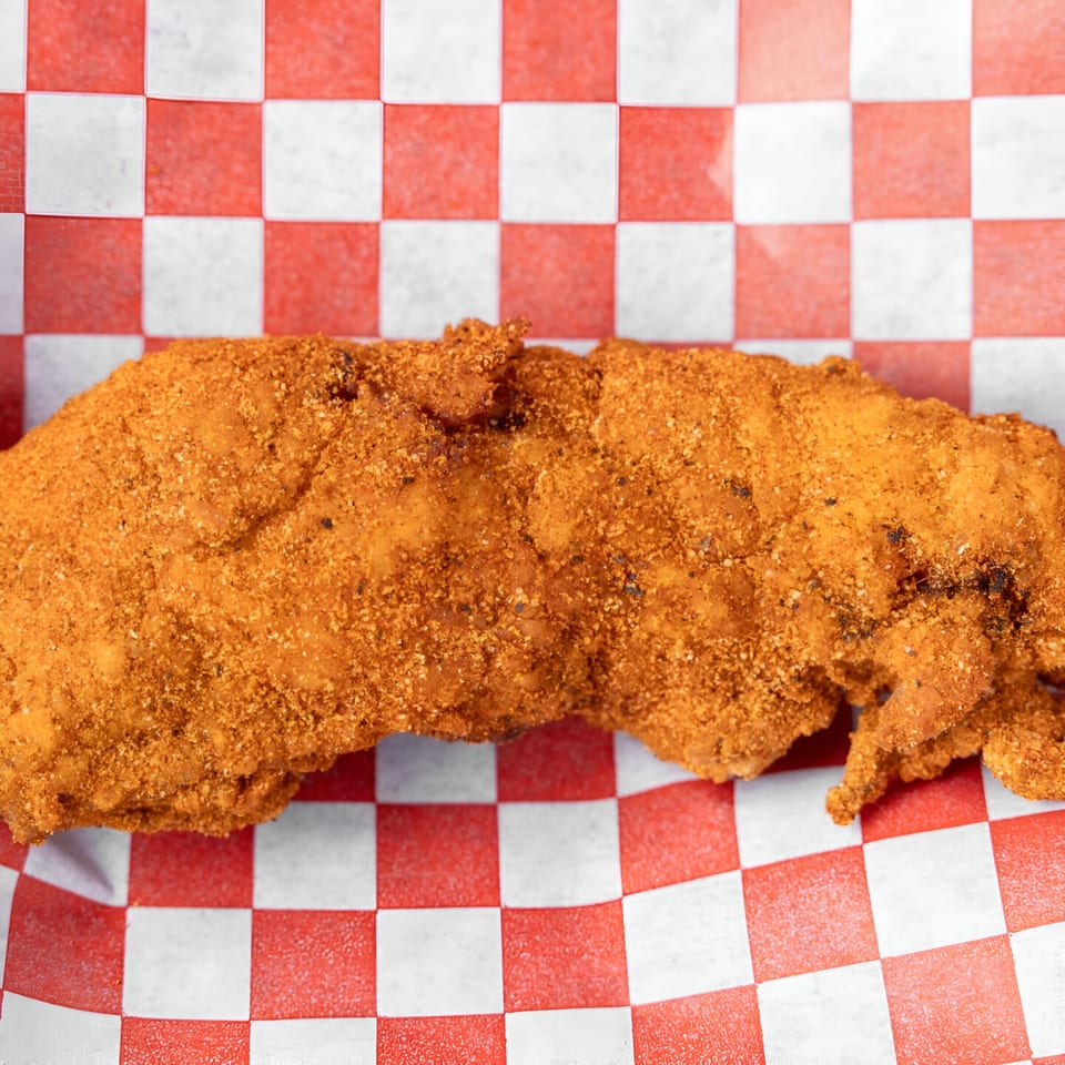 Close-up of Papi's Hot Chicken, a crispy fried chicken fillet covered in spicy seasoning on red and white checkered paper.