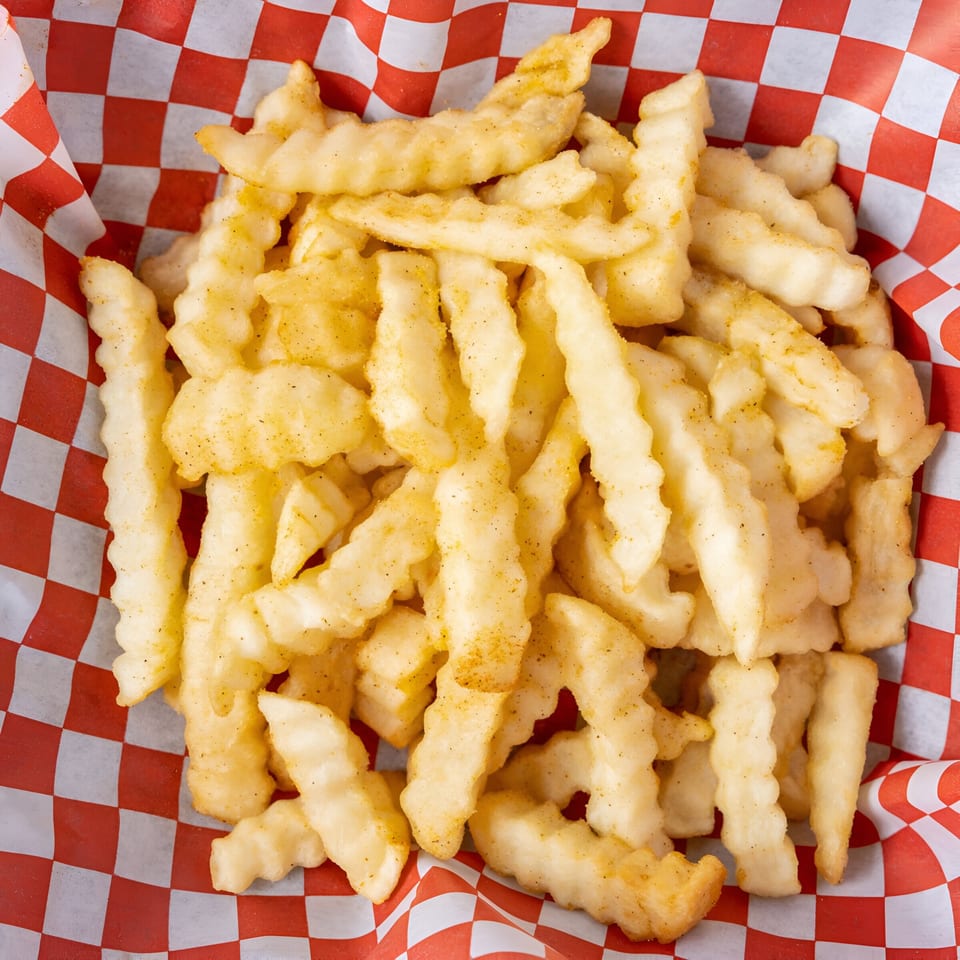A close-up of crinkle-cut fries seasoned with spices, served in a red and white checkered paper basket.
